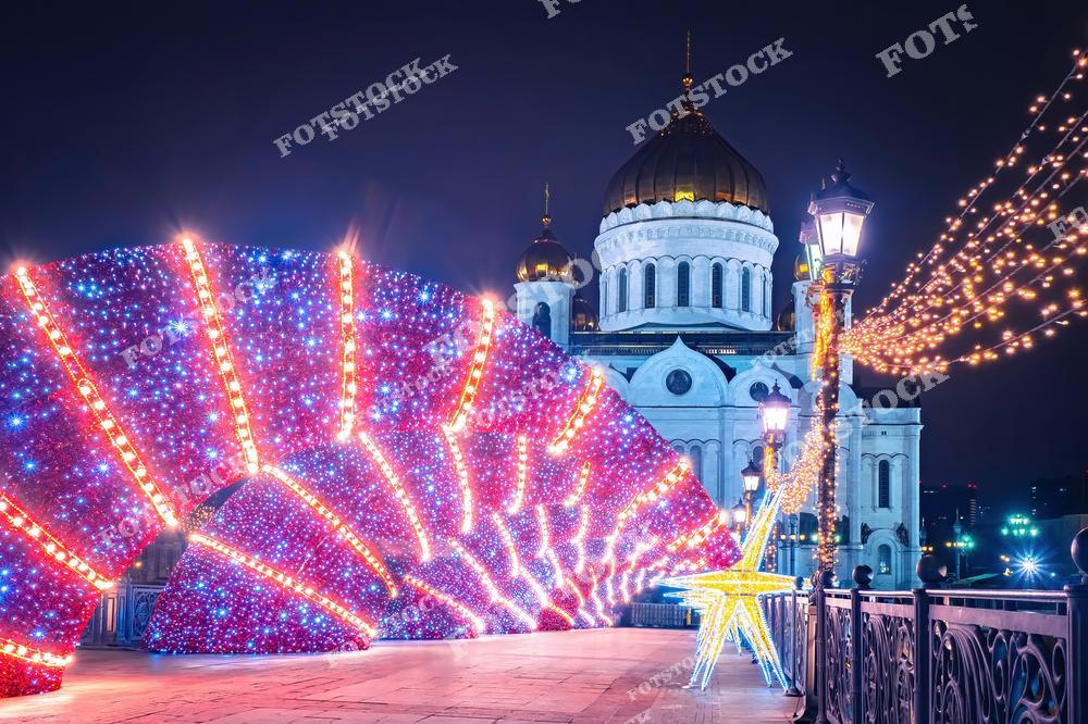 На фотографии изображен Храм Христа Спасителя — главный православный собор Москвы и России. На переднем плане яркая рождественская иллюминация в форме светящейся арки, украшенная гирляндами и звездами. Ночная атмосфера подчеркивает красоту архитектуры храма, купола которого переливаются в свете фонарей. Праздничное настроение усиливают огни гирлянд, украшающих набережную. Эта композиция создаёт уникальное сочетание духовности и новогоднего волшебства.